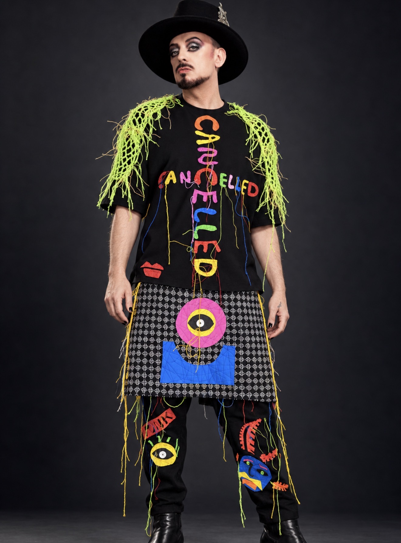 Boy George in a black hat and a colourful outfit with bright green fringe.
